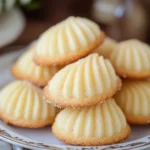 Golden brown Madeleine cookies dusted with powdered sugar in a decorative pan with cream and a whisk nearby.