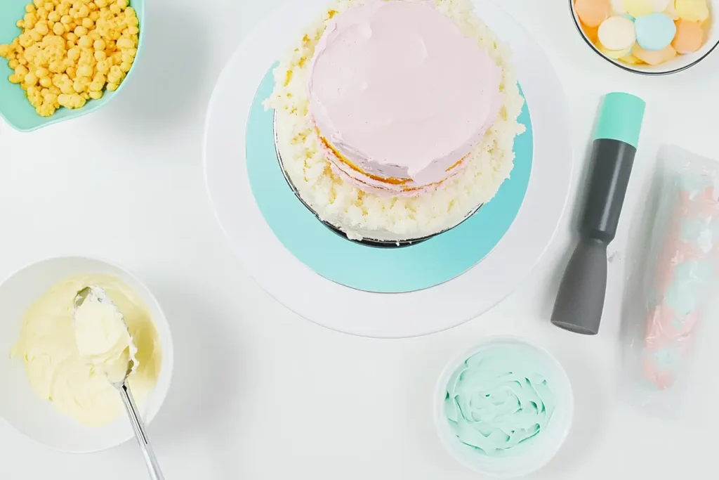 Stacking cake layers with pastel buttercream frosting on a revolving cake stand, surrounded by frosting tools and bowls.