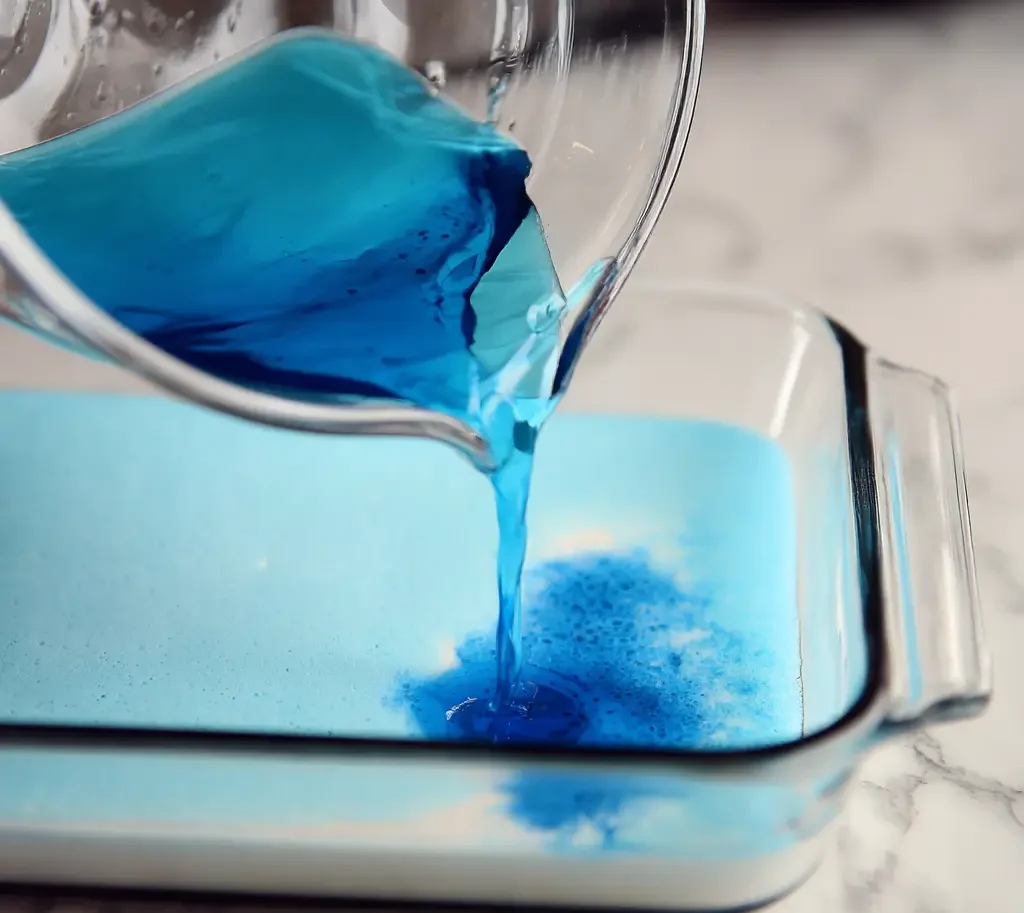 Blue gelatin being poured into glass dish over white layer of jello