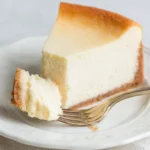 Slice of New York Cheesecake on a white plate with a fork and creamy bite taken out
