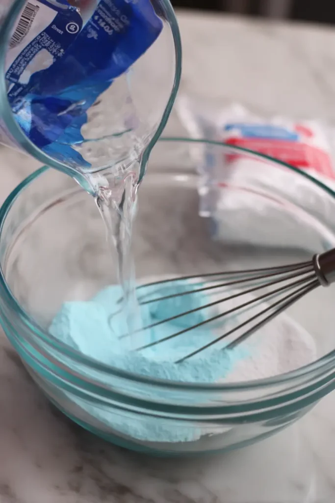 Water being added to blue jello powder in a glass bowl with whisk